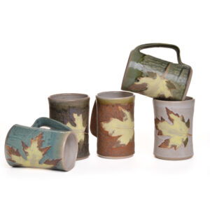 Wheel-thrown mug with leaf decoration. Artist Travis Berning creates his unique, one-of-a-kind mugs on the potter's wheel, then adds a leaf decoration and handle. The mugs are dipped in a wood-ash glaze with white interior.