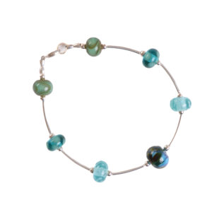 A combination of cool-toned glass beads and sleek-silver metal give this bracelet a fresh and organic feel. Teresa Brittain creates glass beads over a torch, using the centuries-old technique of lampworking, then combines the beads with sterling silver findings and chains to create unique pieces of wearable art.