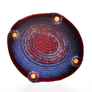 Handmade coiled rope bowl by artist Ann Oakes of North Carolina. Each bowl is a vibrant and unique piece of textile art. The durable rope core is meticulously wrapped in yarn, forming an eye-catching spiral design in complementary shades of rich blue and deep red. Swirls of yarn wrapped around wooden beads add a charming, artisanal touch, making it a perfect functional art piece for any home. Spot clean with a damp cloth or vacuum with a small vacuum cleaner.