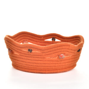 Handmade coiled rope bowl by artist Ann Oakes of North Carolina. Each bowl is a vibrant and unique piece of textile art. The durable rope core is meticulously wrapped in yarn, forming an eye-catching spiral design in shades of orange. Wooden beads around the distinctive, scalloped, bright orange rim add a charming, artisanal touch, making it a perfect functional art piece for any home. Spot clean with a damp cloth or vacuum with a small vacuum cleaner.