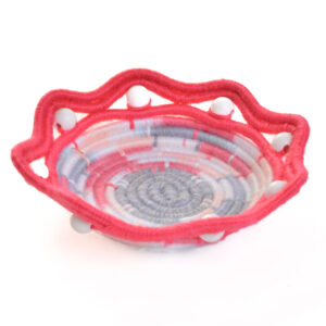 Handmade coiled rope bowl by artist Ann Oakes of North Carolina. Each bowl is a vibrant and unique piece of textile art. The durable rope core is meticulously wrapped in multi-colored yarn, forming an eye-catching spiral design in shades of gray, pink, and off-white. White wooden beads around the distinctive scalloped, bright pink rim add a charming, artisanal touch, making it a perfect functional art piece for any home. Spot clean with damp cloth or vacuum with small vacuum cleaner.