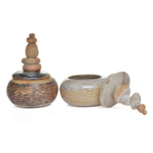 Keep small treasures stored away in this handcrafted stoneware lidded jar featuring a beautifully carved and textured body topped with a stacked-stone lid, creating a balanced presence reminiscent of cairns found in nature, created by North Carolina potter Rebecca Regal. Her ceramics combine hand-building, wheel-throwing, and sculptural techniques, drawing inspiration from Appalachian nature, folklore, and rustic charm. Most of the glazes she uses are original glaze recipes curated by her potter father for over 50 years. Each piece is produced in small batches and is one of a kind. Different colors to choose from.