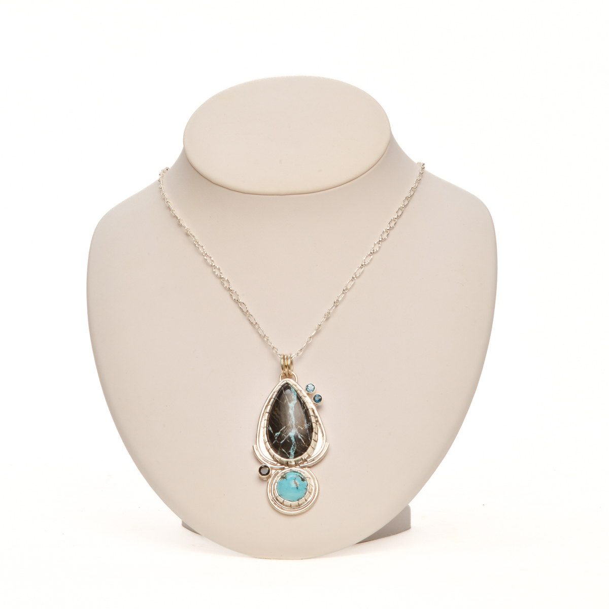 “Blue Lotus” handcrafted sterling silver pendant featuring striking castellated bezel-set Black Jack turquoise accented by a sparkling cluster of faceted black spinel, London blue topaz, and Swiss blue topaz, by Ruthie Cohen of North Carolina.